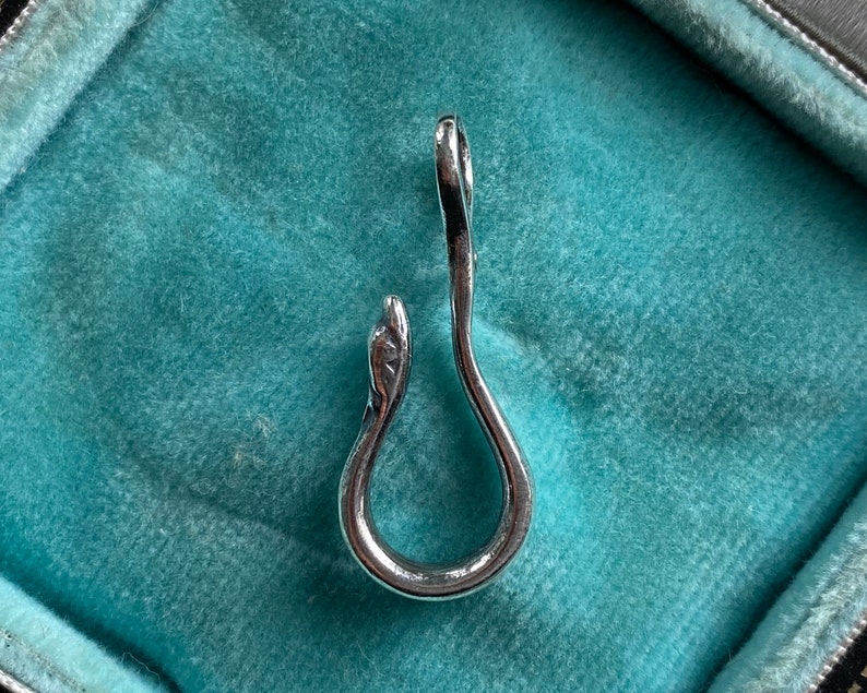 May include: A silver snake-shaped pendant charm with a smooth, polished finish. The charm is in the shape of a snake with its head curled around its body, forming a loop.
