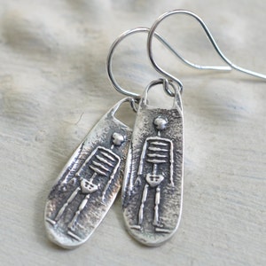 May include: Silver skeleton earrings with a textured finish. The earrings are shaped like a long oval with a skeleton design.