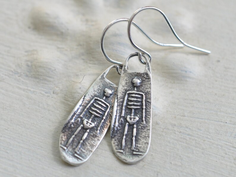 May include: Silver skeleton earrings with a textured finish. The earrings are shaped like a long oval with a skeleton design.