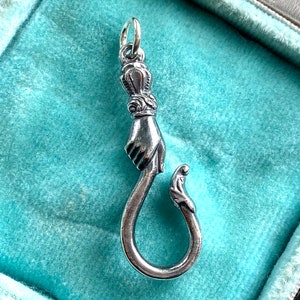 May include: A silver charm in the shape of a hand holding a hook. The hand is detailed with a crown on the top of the finger.