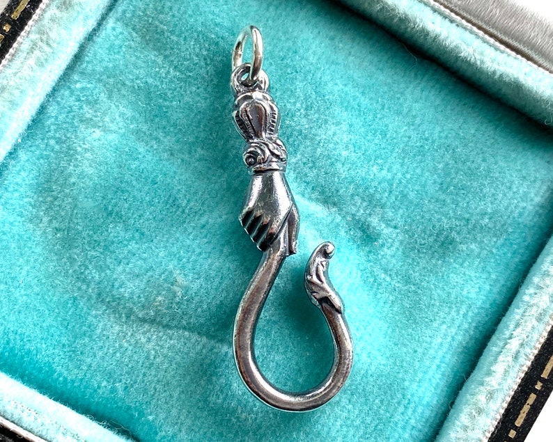 May include: A silver charm in the shape of a hand holding a hook. The hand is detailed with a crown on the top of the finger.