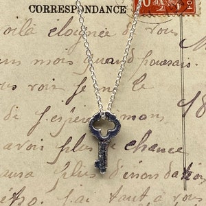 May include: A silver key pendant necklace on a silver chain. The key has an ornate design and is hanging against a vintage piece of paper with French writing.