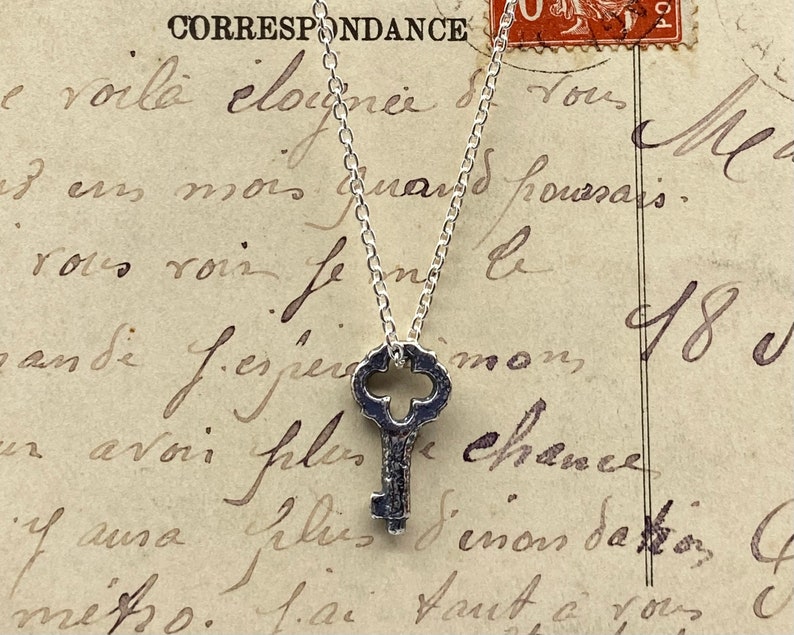 May include: A silver key pendant necklace on a silver chain. The key has an ornate design and is hanging against a vintage piece of paper with French writing.