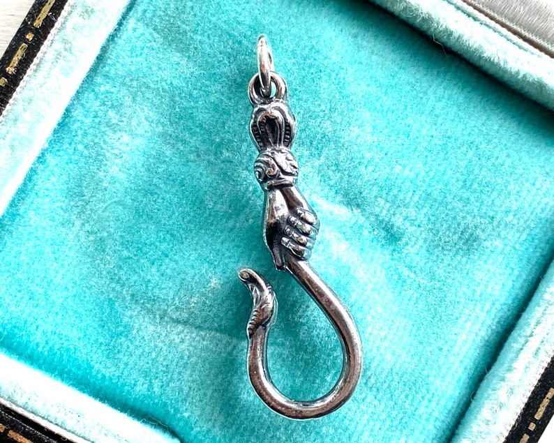 May include: A silver charm in the shape of a hand holding a hook. The charm is detailed with intricate designs and is perfect for adding a unique touch to a necklace or bracelet.