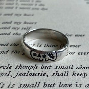 Pu&ograve; includere: Un anello d'argento con un design a forma di teschio. L'anello &egrave; su un libro con testo che recita "This and my heart, This and my self, This and the, is and the, is and the, for ever, er is thine foever, for ever, circle though but small about Devil, jealousie, shall keep out, gift is small but love is all."