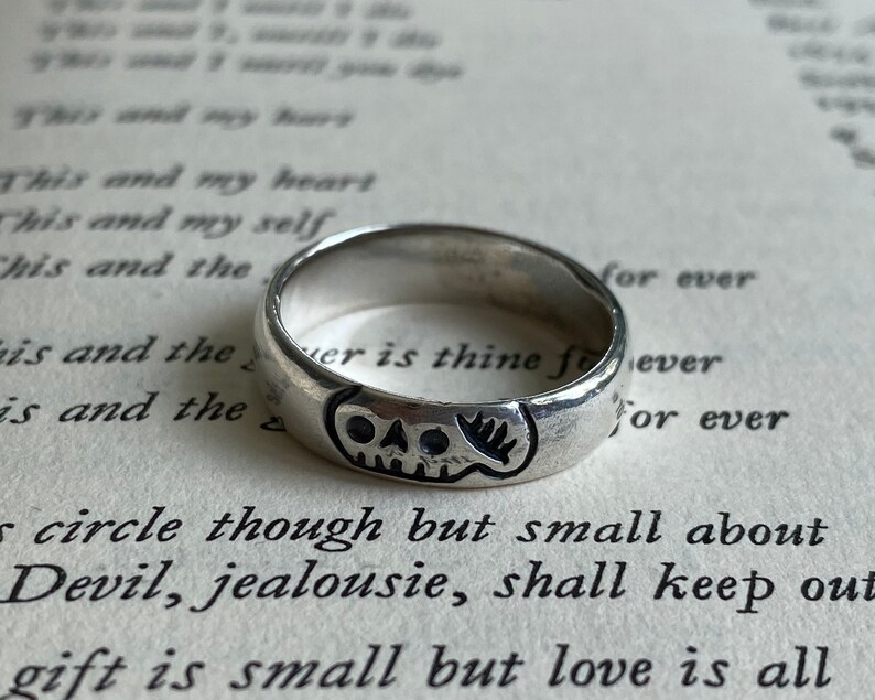 Pu&ograve; includere: Un anello d'argento con un design a forma di teschio. L'anello &egrave; su un libro con testo che recita "This and my heart, This and my self, This and the, is and the, is and the, for ever, er is thine foever, for ever, circle though but small about Devil, jealousie, shall keep out, gift is small but love is all."