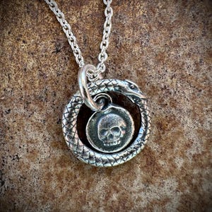 May include: A silver necklace with a pendant featuring a skull surrounded by a snake. The snake is coiled around the skull and its tail is in its mouth.