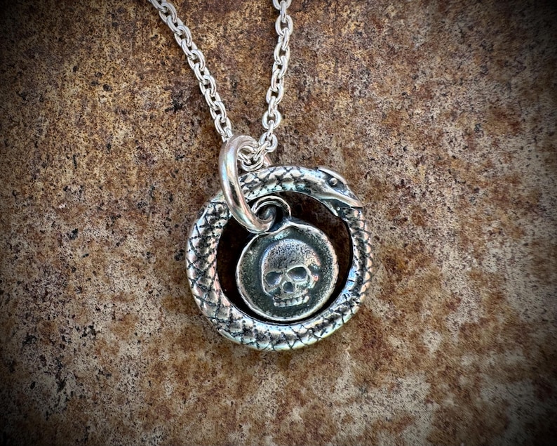 May include: A silver necklace with a pendant featuring a skull surrounded by a snake. The snake is coiled around the skull and its tail is in its mouth.