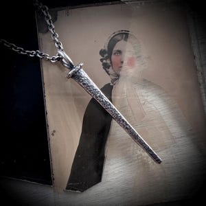 Puede incluir: Un collar con colgante plateado con un colgante en forma de daga con una superficie texturizada. El colgante cuelga de una cadena. El fondo incluye una fotografía vintage de una mujer.