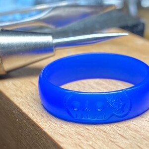 Pu&ograve; includere: Un anello di cera blu con un design a forma di teschio. L'anello &egrave; su una superficie di legno.