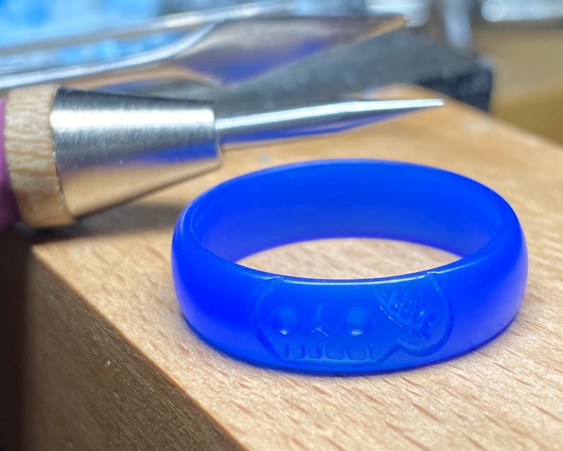 Pu&ograve; includere: Un anello di cera blu con un design a forma di teschio. L'anello &egrave; su una superficie di legno.