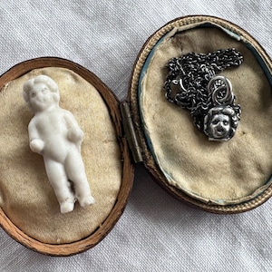 May include: A small porcelain figurine of a child in a vintage wooden box with a silver chain and pendant. The pendant is a face with a detailed design.