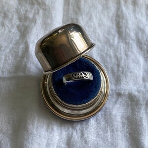 Pu&ograve; includere: Un anello d'argento con un designo a forma di teschio si trova in una scatola per anelli d'argento con un interno in velluto blu.