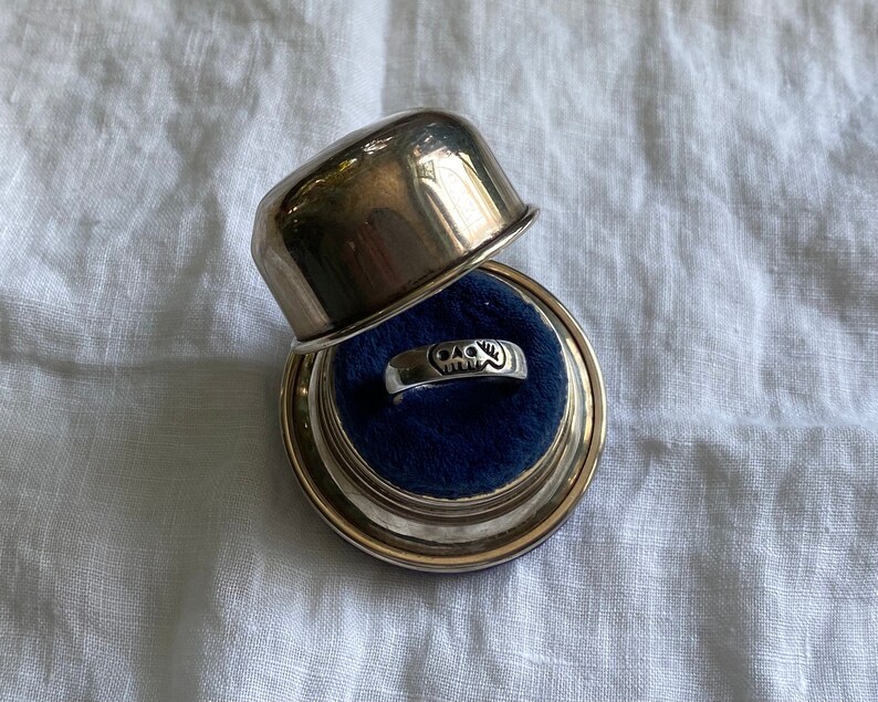 Pu&ograve; includere: Un anello d'argento con un designo a forma di teschio si trova in una scatola per anelli d'argento con un interno in velluto blu.