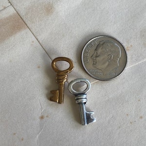 May include: Two small decorative keys, one gold-toned and one silver-toned, next to a U.S. dime. The keys have intricate details and are likely for jewelry or craft projects. The dime is dated 2017.