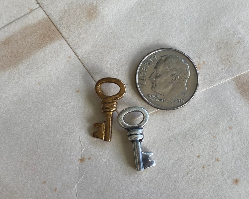 May include: Two small decorative keys, one gold-toned and one silver-toned, next to a U.S. dime. The keys have intricate details and are likely for jewelry or craft projects. The dime is dated 2017.