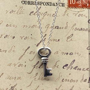 May include: A silver key pendant necklace. The key has a detailed design and hangs from a delicate chain. The background is a vintage postcard with handwritten script and a postage stamp.