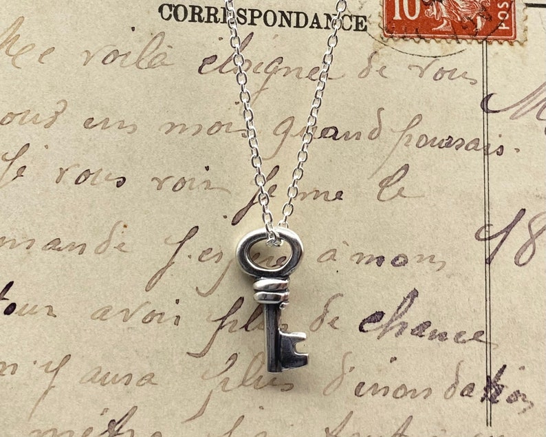 May include: A silver key pendant necklace. The key has a detailed design and hangs from a delicate chain. The background is a vintage postcard with handwritten script and a postage stamp.