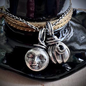 May include: Two silver pendants, one a smiling moon face, the other a stylized pumpkin shape, hang from a silver chain. The pendants rest on a black, glossy surface, with gold and silver chains in the background. The overall theme is celestial and whimsical.