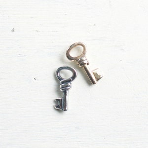 May include: Two antique keys rest on a white, textured surface. One key is silver-toned with a twisted handle, and the other is gold-toned. The keys are likely decorative or vintage items.
