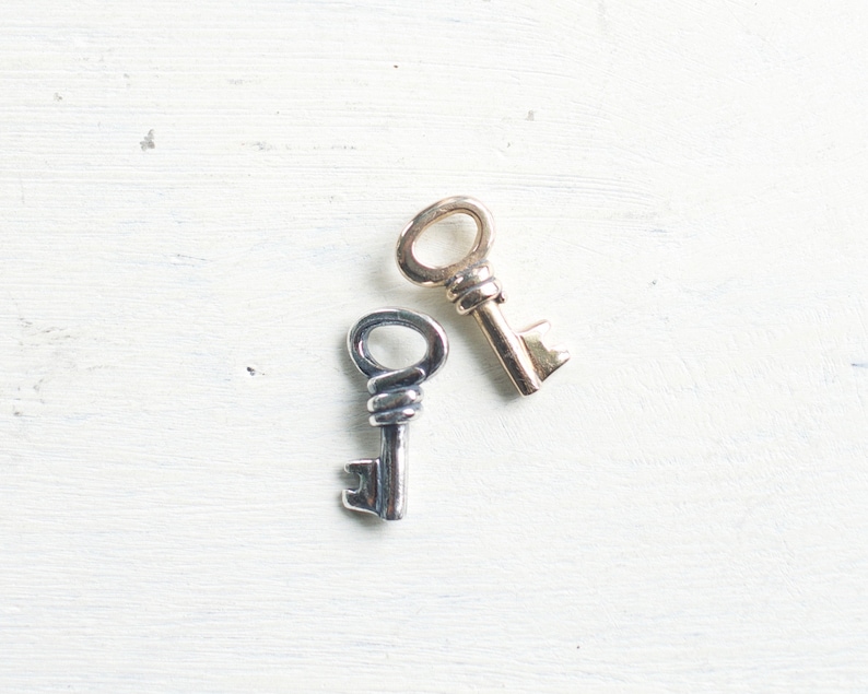 May include: Two antique keys rest on a white, textured surface. One key is silver-toned with a twisted handle, and the other is gold-toned. The keys are likely decorative or vintage items.