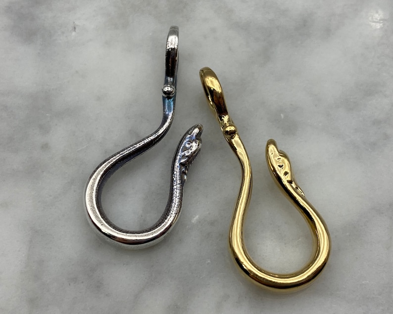 May include: Two silver and gold snake-shaped charms with a loop at the top. The charms are on a white marble surface.