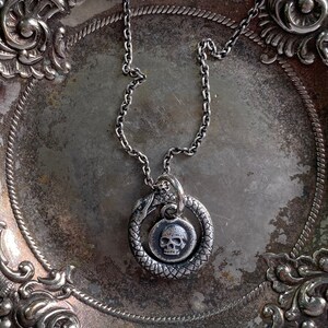 May include: A silver chain necklace with a pendant featuring a skull inside a snake design. The necklace is displayed on a silver tray with ornate detailing.