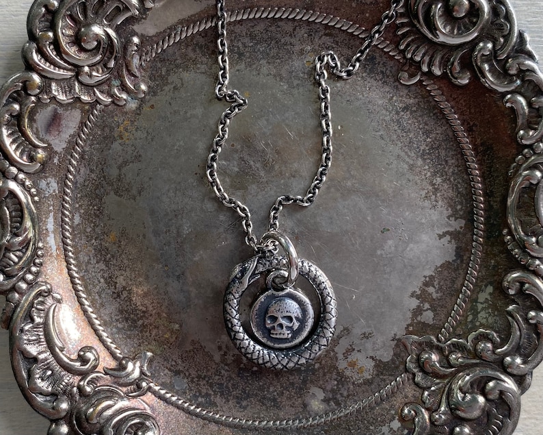 May include: A silver chain necklace with a pendant featuring a skull inside a snake design. The necklace is displayed on a silver tray with ornate detailing.