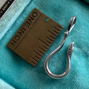 May include: A silver snake-shaped hook with a brass ruler that reads "ONE INCH" in the background.