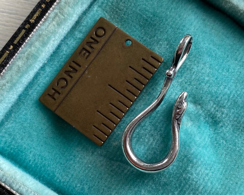 May include: A silver snake-shaped hook with a brass ruler that reads "ONE INCH" in the background.