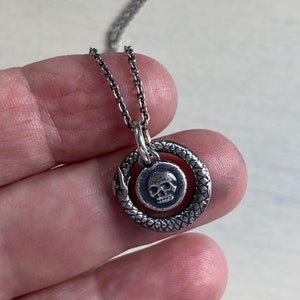 May include: A silver chain necklace with a pendant featuring a skull inside a circle with a snake-like design.