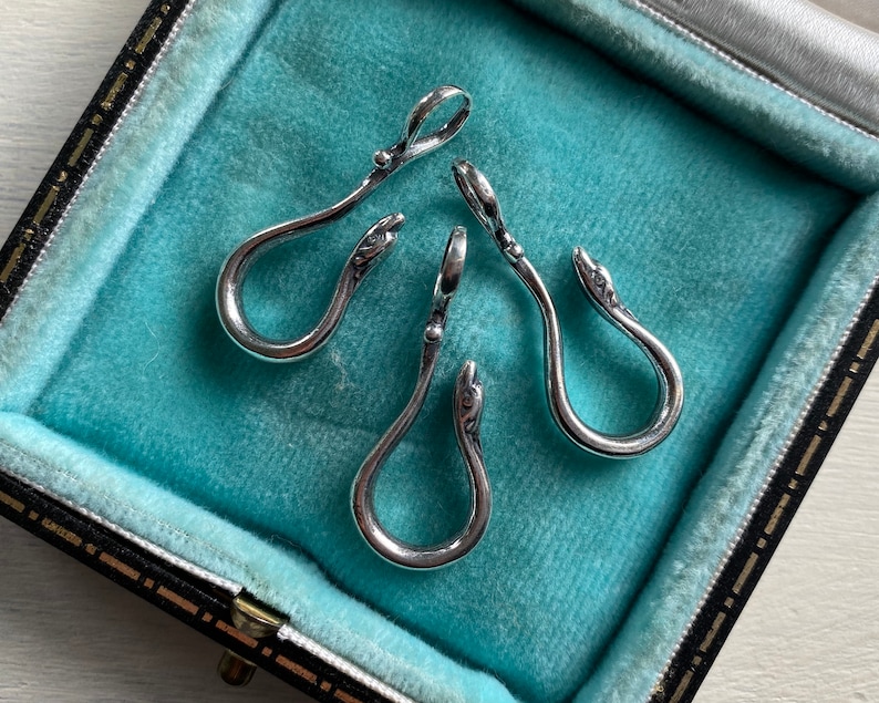 May include: Three silver snake-shaped charms with a hook on the top. The charms are displayed in a blue velvet box.