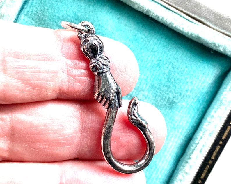 May include: A silver charm in the shape of a hand holding a hook. The charm is detailed with intricate designs and is perfect for adding a unique touch to a necklace or bracelet.