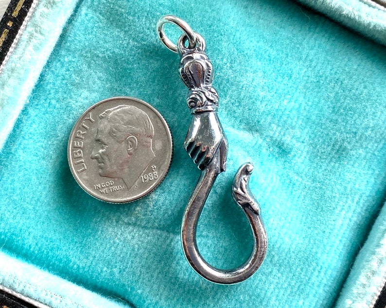 May include: A silver charm in the shape of a hand holding a hook. The charm is detailed with a crown and floral accents. The charm is about the size of a US dime.