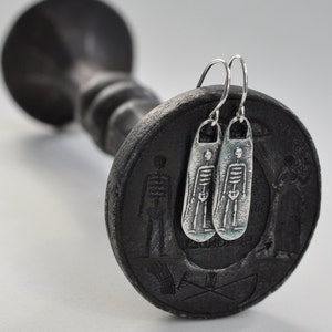 May include: Silver skeleton earrings with a dangling design. The earrings are hanging from a black candlestick.