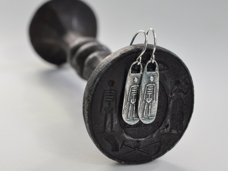 May include: Silver skeleton earrings with a dangling design. The earrings are hanging from a black candlestick.