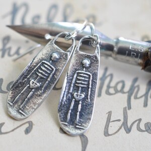 May include: A pair of silver skeleton earrings. Each earring is a long, narrow oval with a textured surface and a raised image of a skeleton with a heart in its chest.