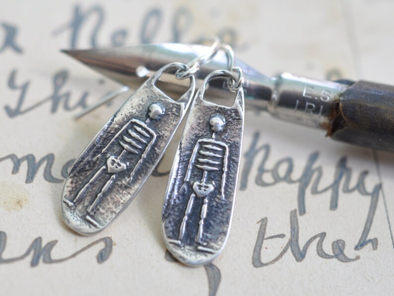 May include: A pair of silver skeleton earrings. Each earring is a long, narrow oval with a textured surface and a raised image of a skeleton with a heart in its chest.