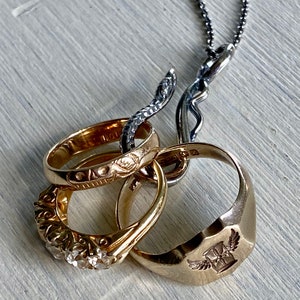 May include: A silver chain necklace with a snake-shaped pendant holding four gold rings. The rings are decorated with intricate designs and one ring has a winged cross emblem.