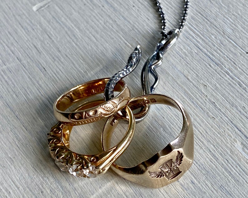 May include: A silver chain necklace with a snake-shaped pendant holding four gold rings. The rings are decorated with intricate designs and one ring has a winged cross emblem.