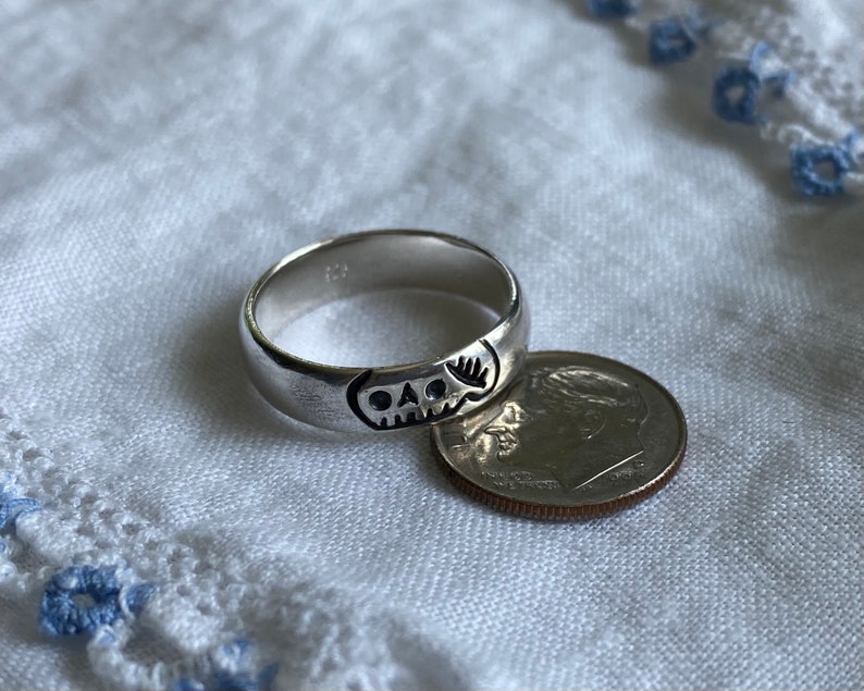 Pu&ograve; includere: Un anello d'argento con un designo a forma di teschio. L'anello &egrave; accanto a una moneta da dieci centesimi di dollaro americano per il confronto delle dimensioni.