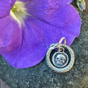 May include: A silver charm featuring a skull inside a snake circle. The charm is on a grey stone background with a purple flower.