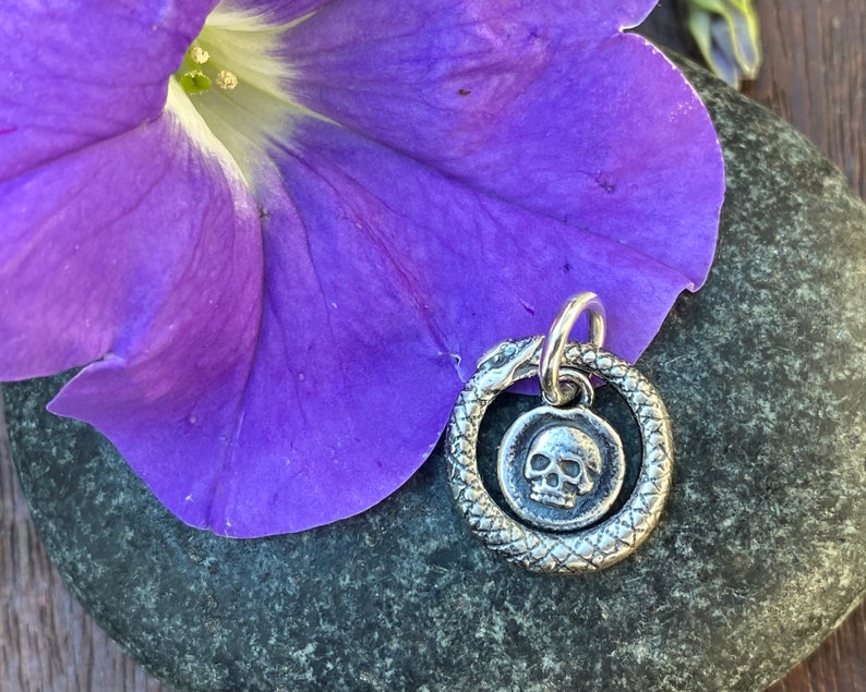 May include: A silver charm featuring a skull inside a snake circle. The charm is on a grey stone background with a purple flower.