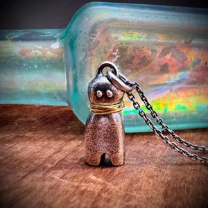May include: A silver-toned metal pendant shaped like a small figure with two circular indentations for eyes, wrapped with thin gold-colored wire around the neck, suspended from a dark silver-toned metal chain link necklace.