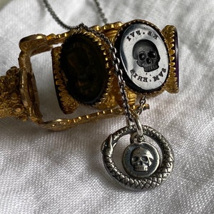 May include: A silver necklace with a skull pendant surrounded by a snake design. The necklace is attached to a gold and purple seal with a skull and the text "M.V.K. 1771" on it.