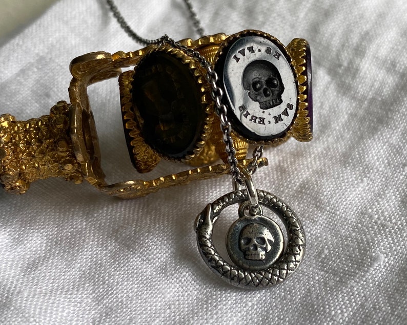 May include: A silver necklace with a skull pendant surrounded by a snake design. The necklace is attached to a gold and purple seal with a skull and the text "M.V.K. 1771" on it.