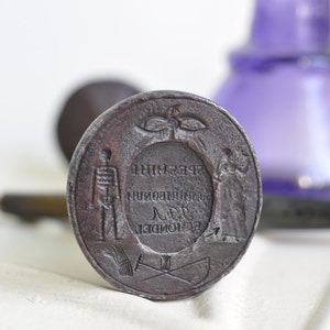 May include: A circular, metal seal with a raised design. The design features a skeleton, a figure holding a staff, and a wreath with a bird inside. The text around the wreath reads "THIM 238", "MUNI", "18", "DE", "LONDON".
