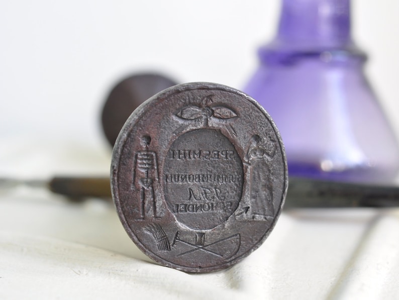 May include: A circular, metal seal with a raised design. The design features a skeleton, a figure holding a staff, and a wreath with a bird inside. The text around the wreath reads "THIM 238", "MUNI", "18", "DE", "LONDON".