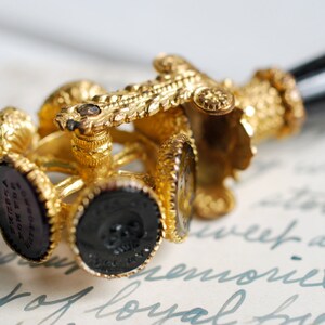 May include: A gold-toned seal with a black handle and multiple interchangeable wax seals. The seals are decorated with intricate designs and feature a black wax impression.