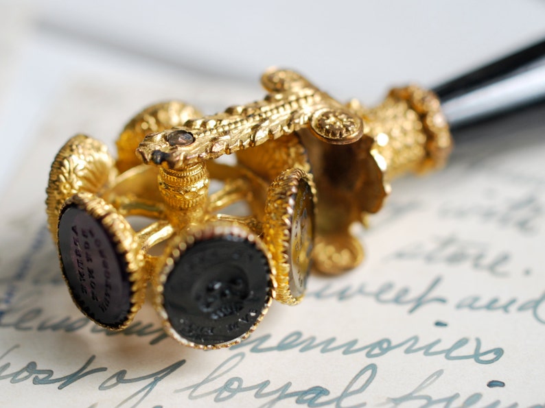 May include: A gold-toned seal with a black handle and multiple interchangeable wax seals. The seals are decorated with intricate designs and feature a black wax impression.